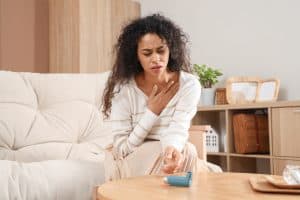 Woman sitting on a couch clutching her chest in discomfort, showing signs of an asthma attack, with an inhaler on the table in front of her.