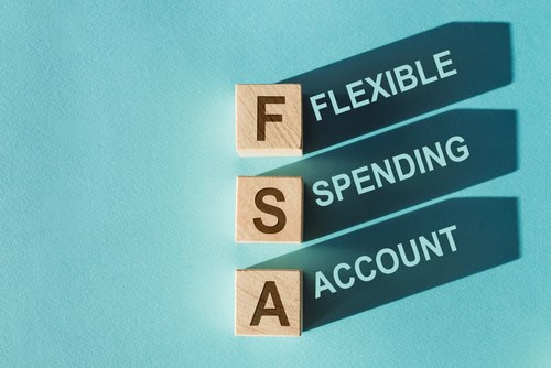 Wooden blocks spelling "FSA" with shadows displaying the words "Flexible," "Spending," and "Account" on a blue background, representing the concept of an FSA (Flexible Spending Account).