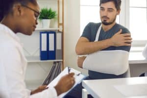 A male patient with a dislocation injury wearing an arm sling sits in a medical office, speaking with a female doctor who is taking notes during the consultation.