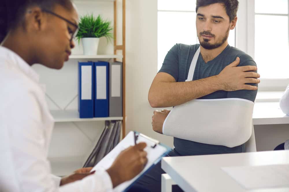 A male patient with a dislocation injury wearing an arm sling sits in a medical office, speaking with a female doctor who is taking notes during the consultation.
