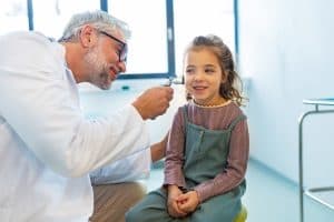 A smiling doctor examines a young girl’s ear with an otoscope in a bright medical office. The girl looks happy and relaxed, wearing a pink and green dress