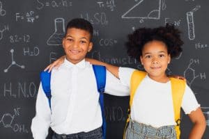 Two smiling children with backpacks standing in front of a chalkboard filled with science and math drawings, symbolizing a back to school theme.