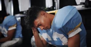 A football player in a blue jersey sits in a locker room with his head in his hand, showing signs of distress or fatigue, possibly suffering from a concussion after a game.
