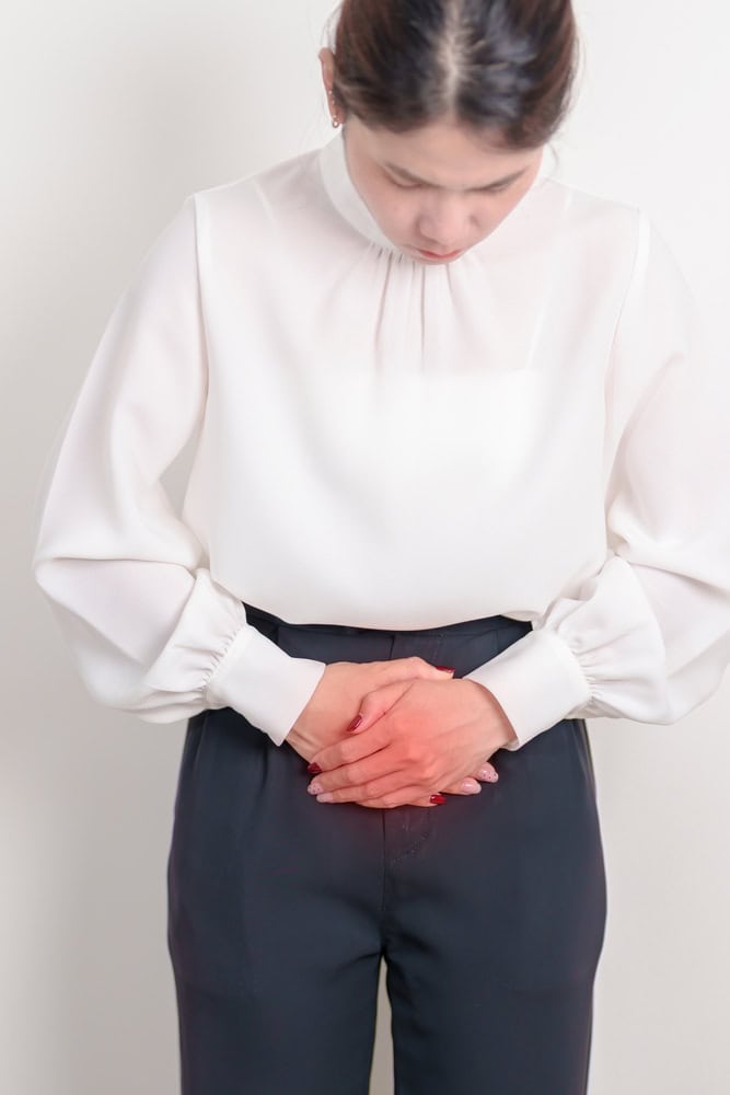 Woman in a white blouse and dark pants holding her lower abdomen in discomfort, possibly indicating symptoms of a UTI (urinary tract infection).